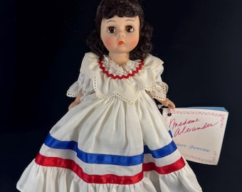 May include: A porcelain doll wearing a white dress with red and blue ribbon accents. The doll has brown hair and brown eyes. The doll is holding a tag that says "Madame Alexander" and "Nature Showcase".
