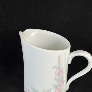 May include: White ceramic creamer with a pink floral design. The creamer has a curved handle and a spout.