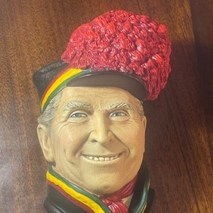 May include: A ceramic bust of a man wearing a black hat with a red feather plume and a red, yellow, and green sash. The man is smiling and has a white beard.