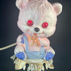 May include: A vintage ceramic nightlight shaped like a pink teddy bear wearing blue overalls. The bear has red eyes and a yellow bow tie. The nightlight sits on a gold-painted metal base.