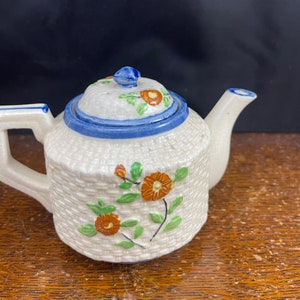 May include: A white ceramic teapot with a woven pattern and orange floral accents. The teapot has a blue rim and a blue knob on the lid.