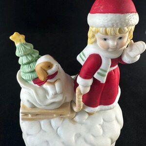 May include: A porcelain figurine of a girl in a red and white winter outfit pulling a sled with a small dog and a Christmas tree on it. The figurine is set against a white snowy background.