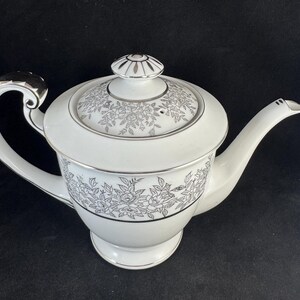 May include: White porcelain teapot with a floral design in black and silver. The teapot has a curved handle, a spout, and a lid with a decorative knob. The floral pattern encircles the body of the teapot, and silver trim accents the edges.