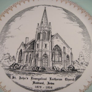May include: A black and white illustration of St. John's Evangelical Lutheran Church in Hubbard, Iowa. The church is a brick building with a tall steeple and a large stained glass window. The image is on a light blue plate with a gold trim.