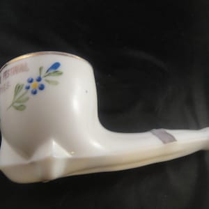 May include: White ceramic pipe with a gold rim and blue and green floral design. The pipe has the text "Festival" and "1968" on the side.