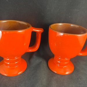 May include: Two orange ceramic mugs with a footed base and a handle. The mugs are empty.