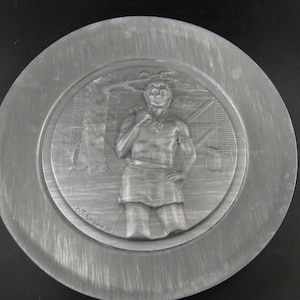 May include: A silver metal plate with a raised design of a man holding a staff. The plate is circular and has a raised rim. The man is wearing a short tunic and has a beard. The design is surrounded by a raised border. The plate is signed "E.J. Brown 79".