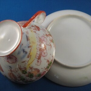 May include: A vintage teacup and saucer set with a floral and geometric design in shades of red, pink, green, and yellow. The cup has a handle and the saucer is plain white.