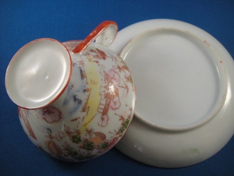 May include: A vintage teacup and saucer set with a floral and geometric design in shades of red, pink, green, and yellow. The cup has a handle and the saucer is plain white.