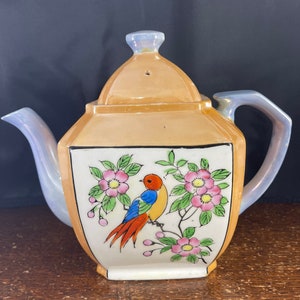 May include: A vintage teapot with a square body and a lid. The teapot is decorated with a colorful bird and floral design on a white background. The teapot is a pale orange color with a pearlescent glaze.