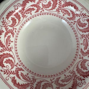 May include: A round white ceramic plate featuring a scalloped edge and a decorative border pattern in red. The border consists of intricate, repeating leaf and vine motifs, framed by a delicate dotted line along the outer rim of the plate.