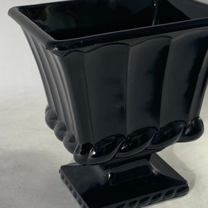 May include: A black glass vase with a square top and a fluted body. The vase has a rope-like design around the base and a square base with a decorative edge.