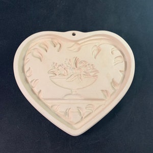 May include: White ceramic heart-shaped cookie cutter with a raised design of a bowl of flowers. The cutter has a hole at the top for hanging.