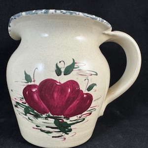 May include: Cream-colored ceramic pitcher with a large handle and a spout. The pitcher is decorated with three red apples and green leaves. The rim has a blue and white speckled design.