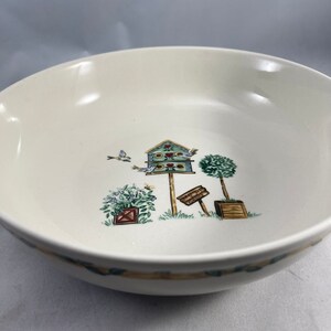 May include: A white ceramic bowl with a green, blue, and brown birdhouse design. The bowl has a rim with a green and brown pattern.