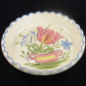 May include: A cream-colored ceramic pie plate with a scalloped edge, painted with a floral design. The flowers are pink and blue, with green leaves and a pink pot. The rim is trimmed with a blue wavy line.