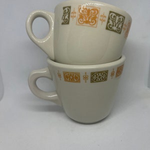 May include: Two white ceramic coffee mugs with a brown and green geometric pattern around the top edge. The mugs are stacked on top of each other.