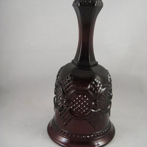 May include: A dark burgundy glass bell with intricate embossed floral and geometric patterns. The bell has a long neck and a textured top. The base is round and the bell is made of a shiny, reflective material.