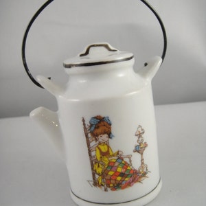 May include: White porcelain milk can with a black wire handle. The can has a colorful illustration of a girl sitting in a rocking chair, sewing a quilt.