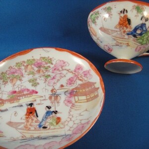 May include: A white porcelain teacup and saucer set with a hand-painted design of Japanese women in a boat with cherry blossoms and a pagoda. The cup has a rounded shape and a handle. The saucer has a scalloped edge.
