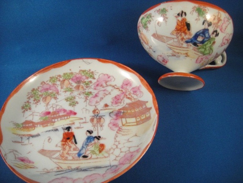 May include: A white porcelain teacup and saucer set with a hand-painted design of Japanese women in a boat with cherry blossoms and a pagoda. The cup has a rounded shape and a handle. The saucer has a scalloped edge.