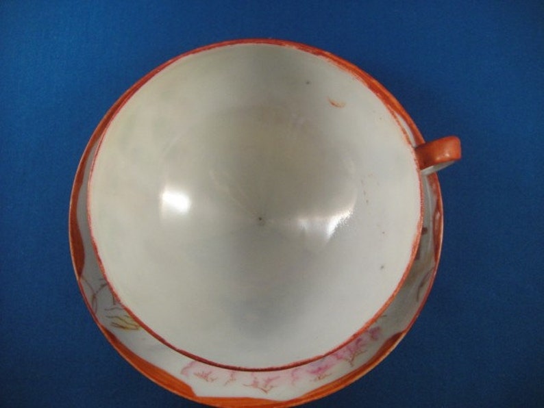 May include: A white teacup and saucer with a floral design in shades of pink and orange. The saucer has a scalloped edge.