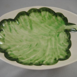 May include: A white ceramic dish with a green leaf design. The leaf is painted in shades of green and has a slightly textured surface.