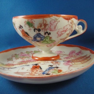 May include: A white porcelain teacup and saucer set with a floral and geisha design. The cup has a handle and a wide, shallow bowl. The saucer has a scalloped edge.