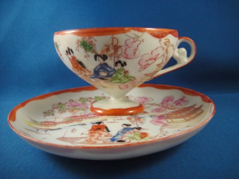 May include: A white porcelain teacup and saucer set with a floral and geisha design. The cup has a handle and a wide, shallow bowl. The saucer has a scalloped edge.