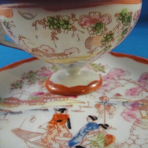 May include: A white porcelain teacup and saucer set with a floral design and a scene of Japanese figures in a landscape. The cup has a handle and a wide, rounded shape. The saucer has a scalloped edge.