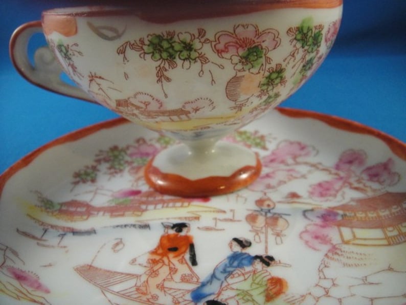 May include: A white porcelain teacup and saucer set with a floral design and a scene of Japanese figures in a landscape. The cup has a handle and a wide, rounded shape. The saucer has a scalloped edge.