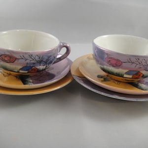 May include: Two matching teacups and saucers with a purple, pink, and yellow floral design. The cups and saucers feature a painted scene of a house with a tree and a pink flower.