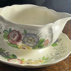 May include: A white ceramic gravy boat with a floral design. The boat has a scalloped edge and is decorated with pink, purple, yellow, and green flowers. It is sitting on a matching saucer.