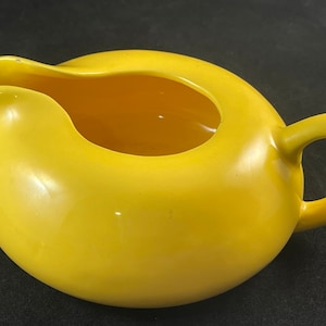 May include: A bright yellow ceramic creamer or gravy boat with a rounded body and a handle. The creamer is empty and ready to be filled.