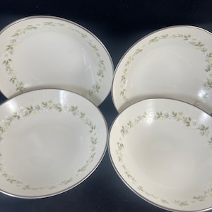 May include: Four off-white ceramic bowls with a silver rim and a floral design. The floral design features white flowers, green leaves, and yellow accents. The bowls are arranged on a black surface.