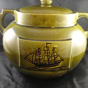 May include: A green ceramic crock with a lid and two handles. The crock has a black silhouette of a sailing ship on the front.