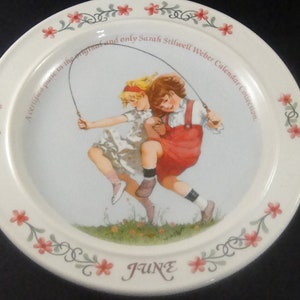 May include: A white ceramic plate with a floral border and a picture of two girls jumping rope. The plate says "A carefree time in the springtime" and "and only Sarah Stilwell Weber Calendar Collection" around the picture. The bottom of the plate says "JUNE".