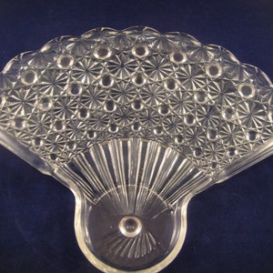 May include: A clear glass fan-shaped dish with a decorative pattern of small circles and stars. The dish has a scalloped edge and a raised center.