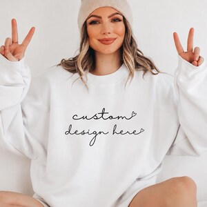 Può includere: Felpa bianca con la scritta "custom design here" in nero. La felpa ha maniche lunghe e girocollo. Un berretto beige è indossato sulla testa.