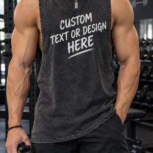 May include: A charcoal grey acid-wash sleeveless tank top featuring the white printed text CUSTOM TEXT OR DESIGN HERE on the chest. The garment has a crew neckline and wide arm openings, showcasing a distressed fabric texture throughout the piece.