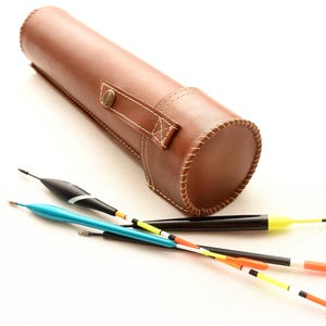 May include: A brown leather fishing float case with a button closure and white stitching. Several colorful fishing floats with orange, yellow, black, and blue accents are arranged near the case.
