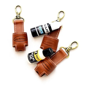 Könnte beinhalten: Drei braune Leder-Schlüsselanhänger mit einem Metallclip und einer Schlaufe zum Halten kleiner Flaschen. Die Schlüsselanhänger sind mit "Toby Machenwirth" beschriftet. Die Flaschen sind mit "DUCK", "GULFF" und "loon ROYAL GEL" beschriftet.