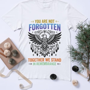 Può includere: Una t-shirt bianca a maniche corte con la grafica di un'aquila nera con motivi geometrici e piume. Il testo recita YOU ARE NOT FORGOTTEN in blu e viola, e TOGETHER WE STAND IN REMEMBRANCE in arancione e verde chiaro.
