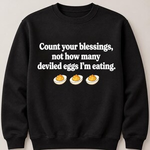 May include: A black crewneck sweatshirt featuring the white text "Count your blessings, not how many deviled eggs I'm eating." centered on the chest, accompanied by a graphic illustration of three halved deviled eggs with yellow yolks.