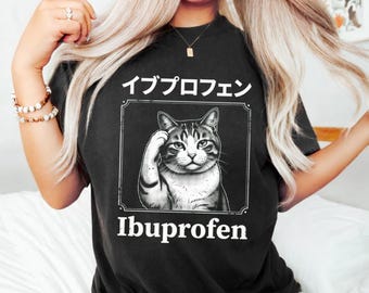 May include: A black t-shirt featuring a graphic of a cat with one paw raised to its head. Above the cat is the Japanese text "イブプロフェン" and below the cat is the English text "Ibuprofen". The design is enclosed in a rectangular frame.