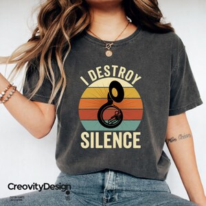 Może przedstawiać: Ciemnoszary t-shirt z grafiką czarnego suzafonu umieszczoną na tle wzoru retro zachodu słońca z poziomymi pasami w kolorze żółtym, pomarańczowym i turkusowym. Napis I DESTROY SILENCE jest nadrukowany jasną żółtą czcionką o pogrubionym kroju.