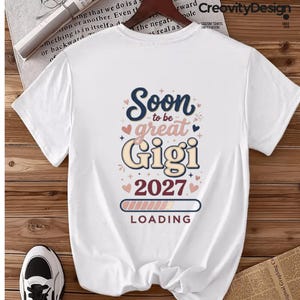May include: A white short-sleeved t-shirt featuring a graphic print that reads Soon to be great Gigi 2027 LOADING. The text is displayed in navy blue, pink, and cream colors, accented with small heart and star icons and a progress bar graphic.