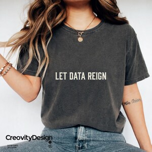 May include: A charcoal grey short-sleeve t-shirt featuring the text LET DATA REIGN printed in a light cream colour across the chest. The shirt has a crew neckline and a washed, vintage-inspired fabric texture, paired with blue denim jeans.