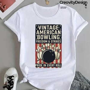 May include: A white short-sleeve t-shirt featuring a rectangular graphic with the text VINTAGE AMERICAN BOWLING FREEDOM & STRIKES PRIDE IN EVERY ROLL. The design includes a black bowling ball and white pins over a red, white, and blue striped background.