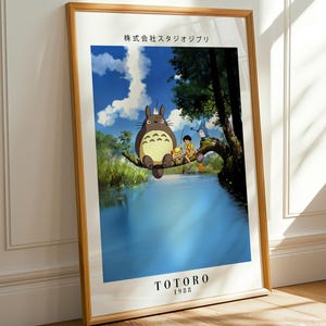 May include: Framed poster featuring a scene from the animated film "My Neighbor Totoro." The artwork depicts Totoro and other characters near a body of water. The poster has a wooden frame and is leaning against a white wall and wooden floor.
